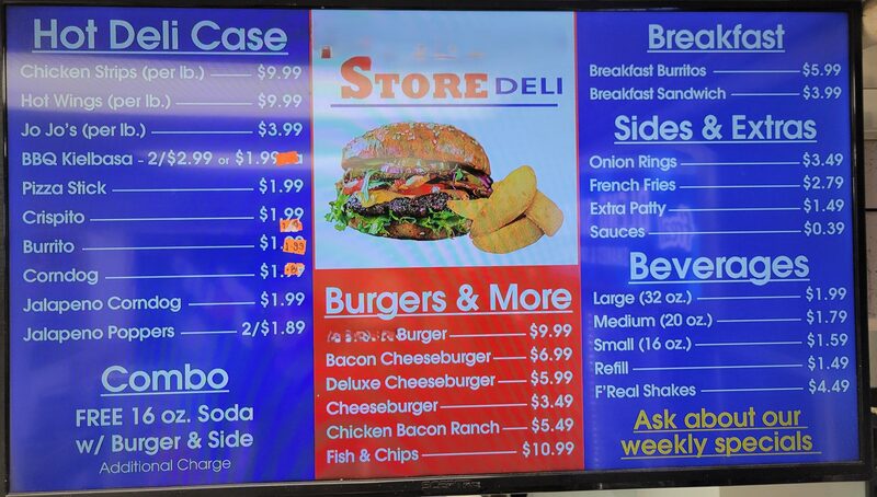 A gas station deli menu displayed on a digital screen, with orange price gun stickers stuck directly over some of the digital prices
